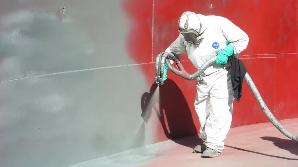 Worker in a white protective suit and respirator spray-painting a red metal surface grey.