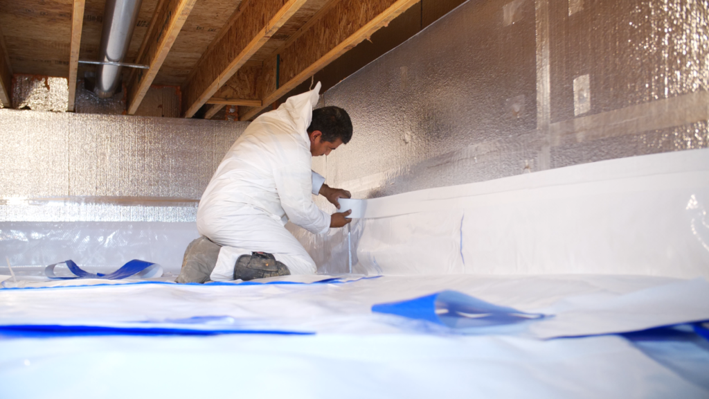 A worker in a white protective suit applies white tape to silver insulation in a construction site.