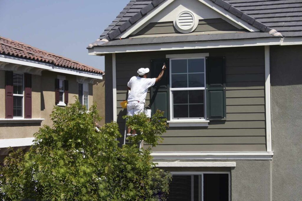 A painter in white clothing on a ladder paints dark green shutters on the second story of a house.