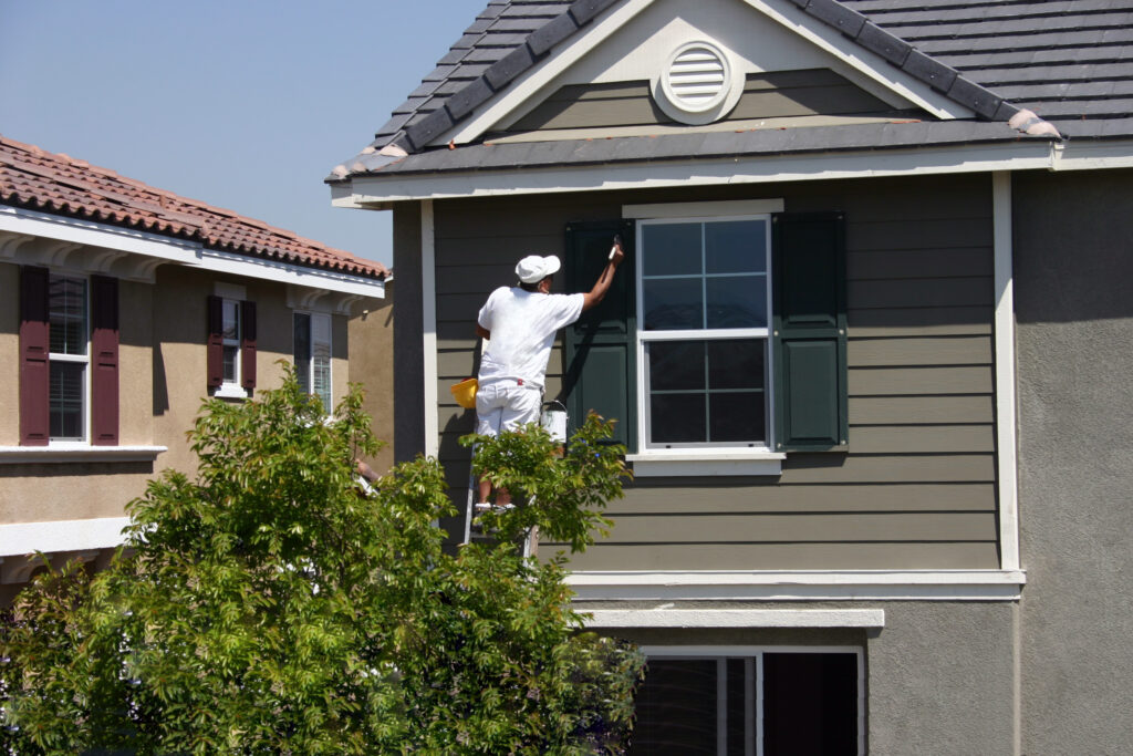 Painter in white clothing on a ladder painting dark green window shutters on a house.