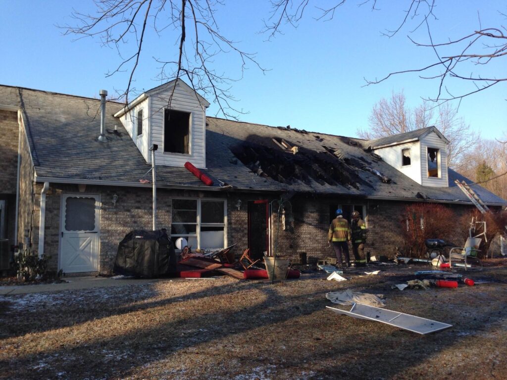 Fire-damaged brick house with a collapsed roof and two firefighters standing amidst debris on the lawn.