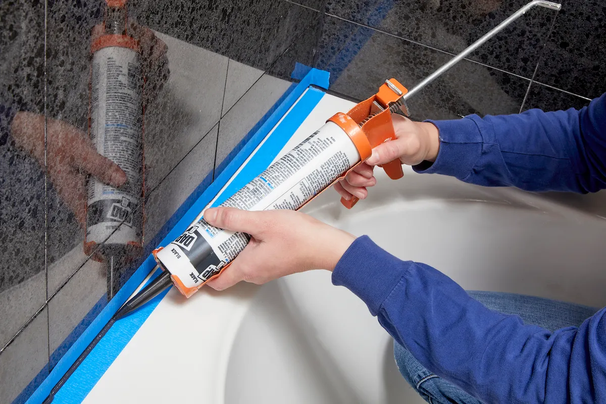Applying black silicone caulk to a bathtub joint using a caulking gun and painter's tape.