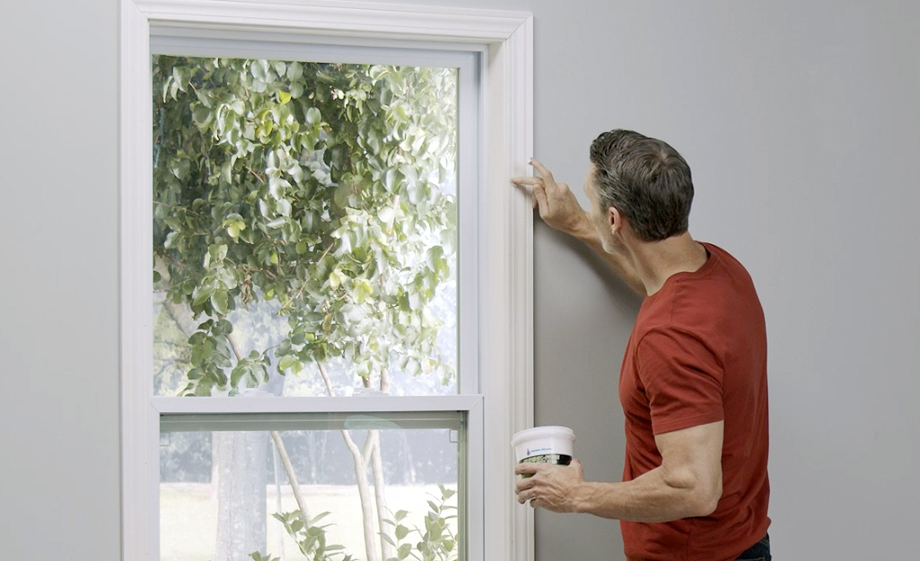 A man in a red t-shirt applies white caulk to the trim of a window frame.
