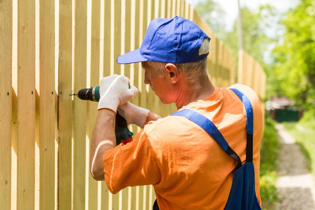 A construction worker wearing a blue cap and orange shirt uses a power drill to attach a board to a wooden fence.
