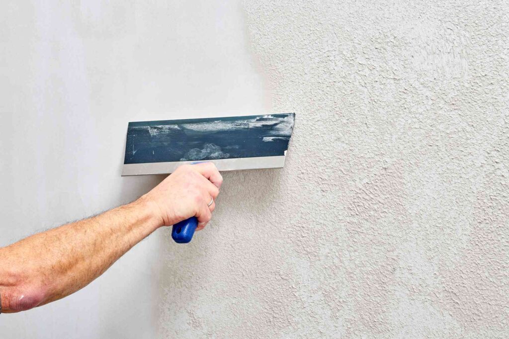 Hand using a taping knife to apply smooth joint compound over a textured wall surface.
