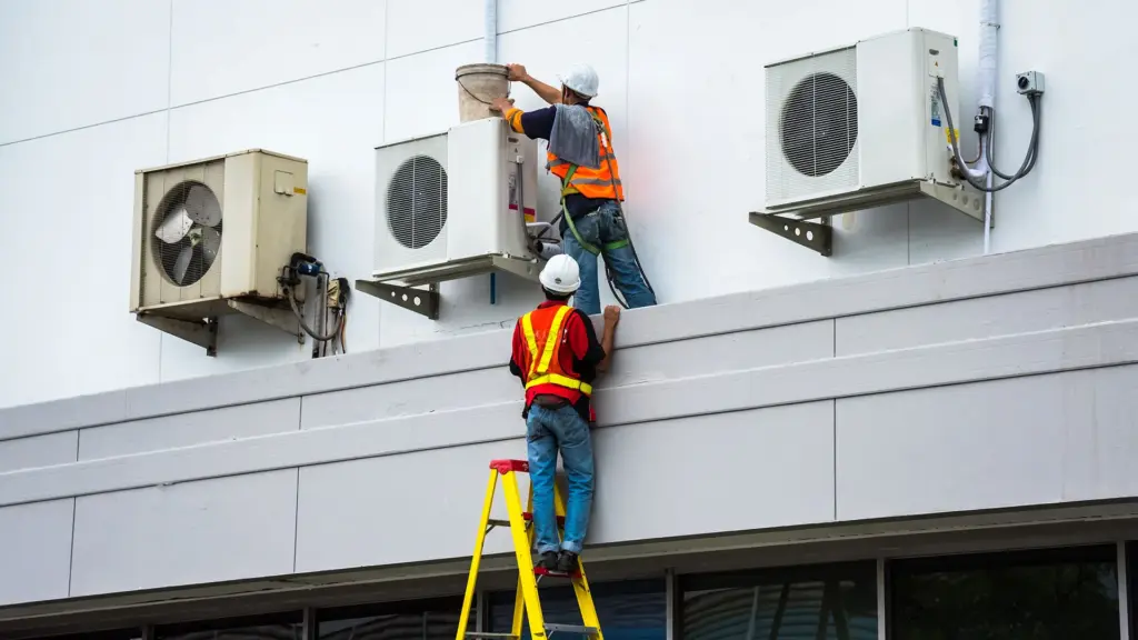 Two technicians in high-visibility vests and hard hats servicing AC units on a building wall.
