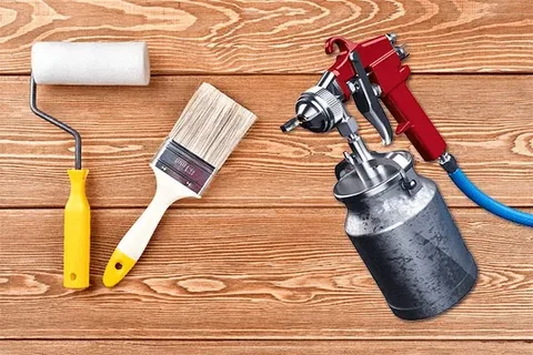 Paint roller, paintbrush, and paint sprayer on a wooden surface.