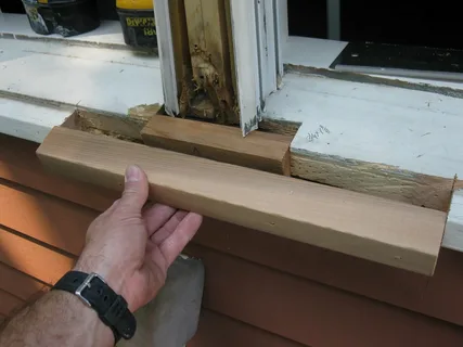 Hand holding a new wooden trim piece against a damaged white window sill for repair.