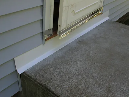 Damaged bottom edge of a white door and frame next to grey house siding on a concrete porch.