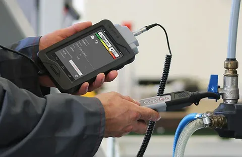 Technician using a handheld diagnostic device and probe to test an industrial valve and piping system.