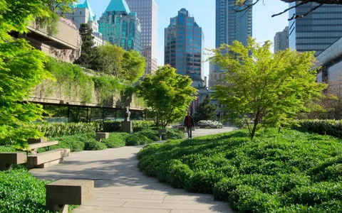 Winding stone path through a lush urban park with concrete benches and city skyscrapers in the background.