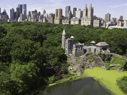 Belvedere Castle in Central Park, surrounded by lush greenery and a pond, with the NYC skyline in the background.