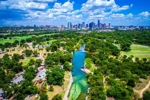 Turquoise river winding through a lush green park leading toward a distant city skyline under a bright, cloudy sky.