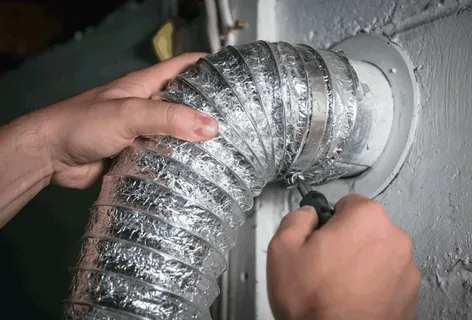 Hands securing a flexible silver foil vent duct to a wall opening with a screwdriver.