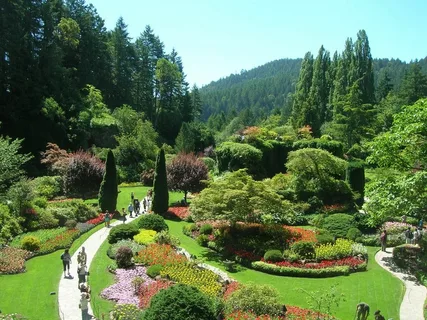 Lush landscaped garden with winding paths, vibrant flower beds, and visitors against a forested mountain backdrop.