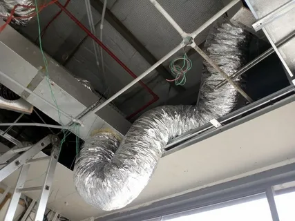 Flexible silver foil air duct connected to a ceiling HVAC system in an unfinished room.