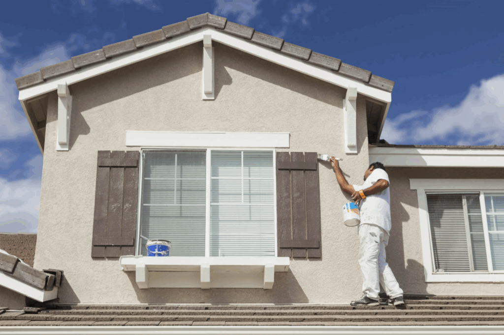 A house painter in white stands on a roof, painting the trim of a window shutter on a beige house.