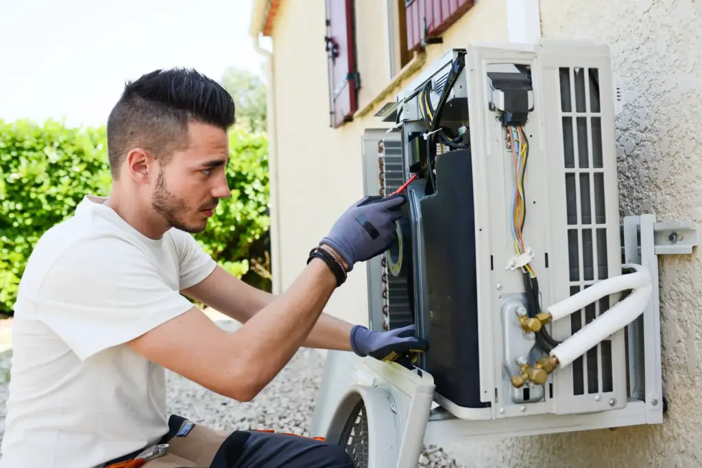 Technician wearing gloves repairing an outdoor air conditioning unit mounted on a house wall.