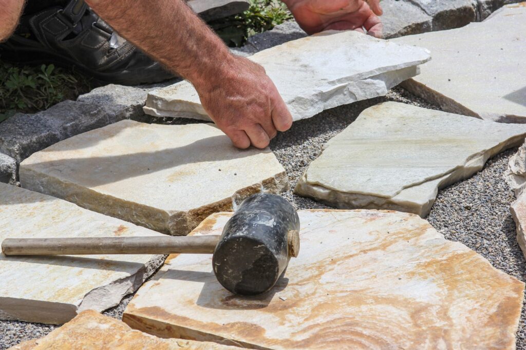 A mason's hand places a flagstone onto a gravel bed, with a rubber mallet resting on the path nearby.
