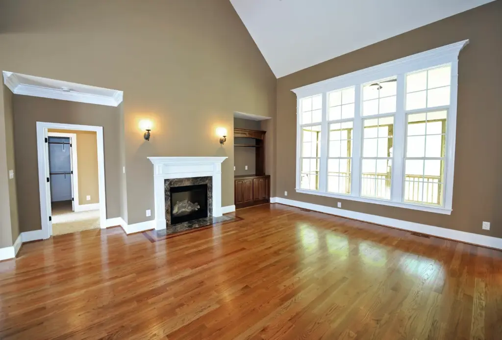 Spacious empty living room with hardwood floors, vaulted ceiling, white mantle fireplace, and large windows.
