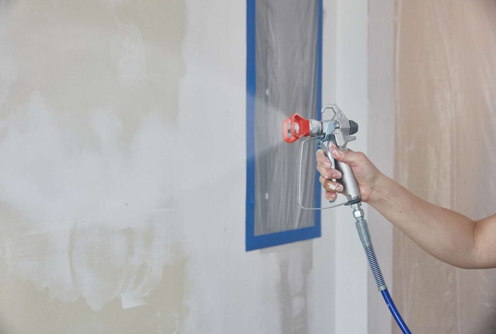 A hand uses a paint spray gun to paint a white wall, with a blue-taped area protected by plastic.