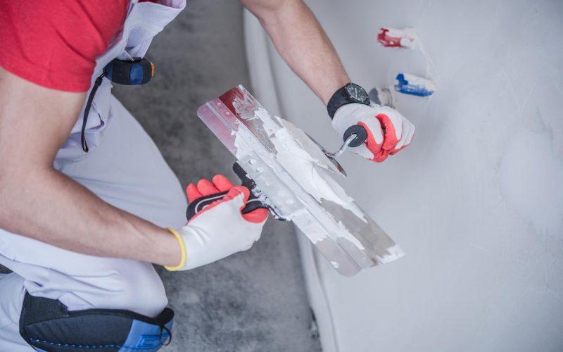 A construction worker using a wide trowel to apply plaster or joint compound to a white wall.