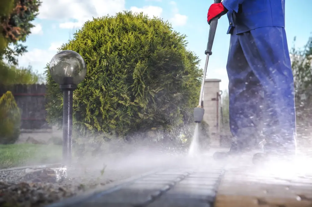 A person in blue coveralls uses a pressure washer to clean a stone walkway in a garden.