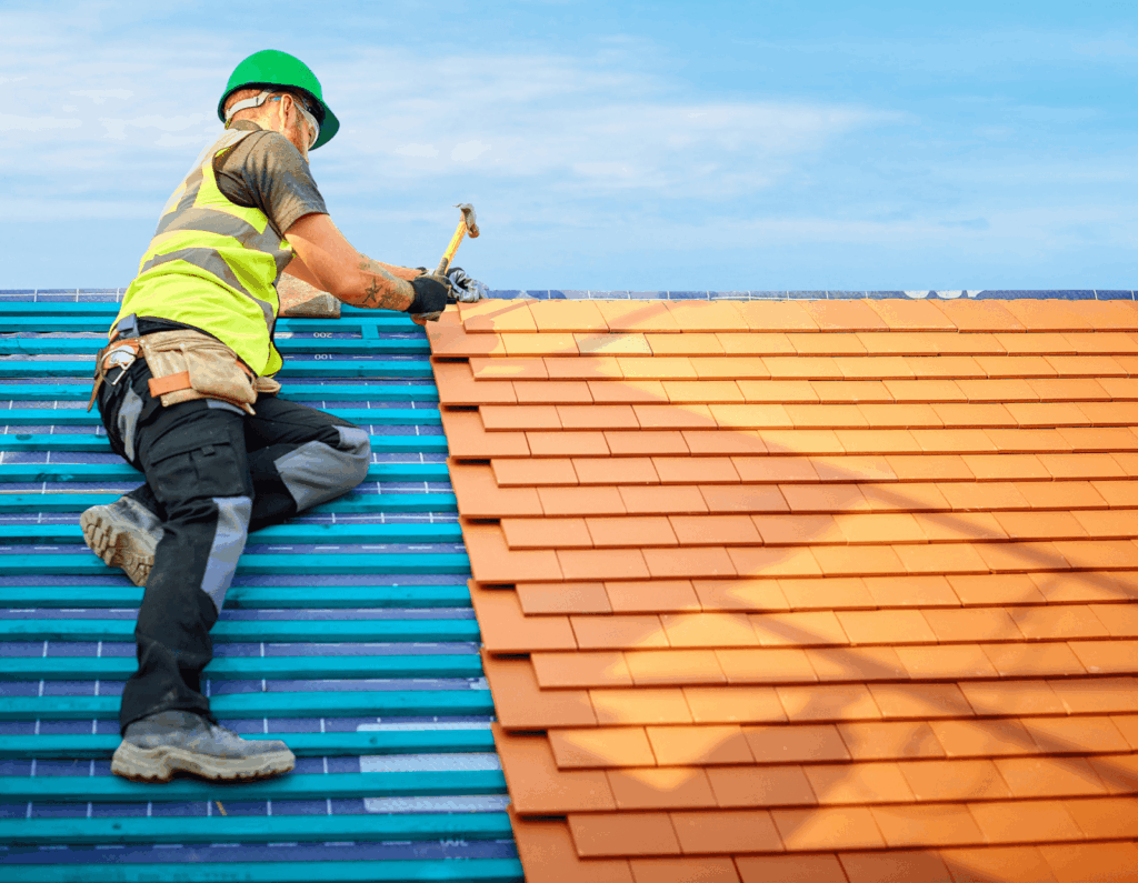 Construction worker in a green hard hat and safety vest installs orange roof tiles.
