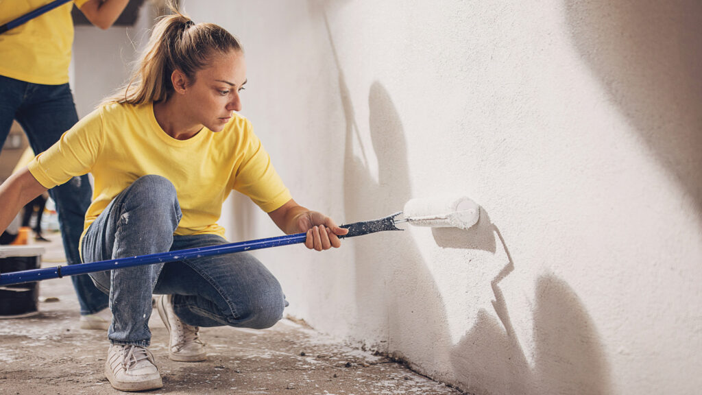 A woman in a yellow t-shirt squats to paint a white wall with a paint roller.