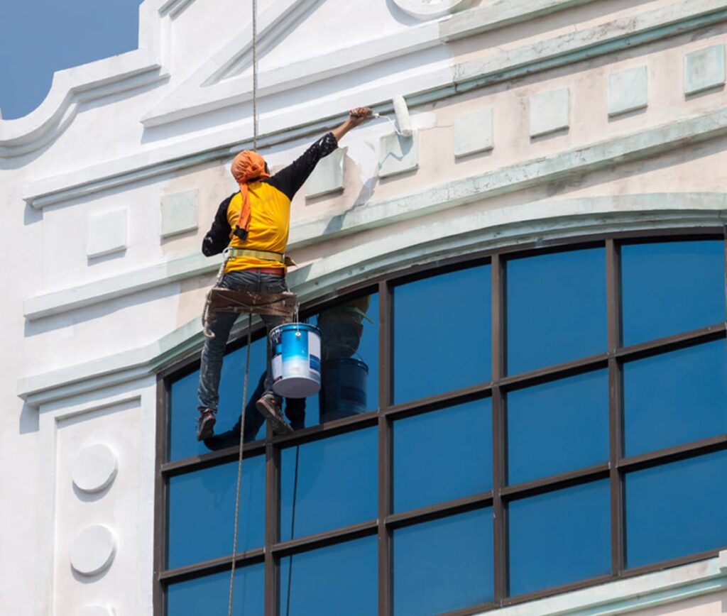 Painter suspended by safety ropes paints the white exterior facade of a building above large blue windows.