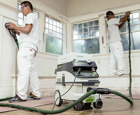 Two workers sanding walls and windows using power tools connected to a large industrial vacuum cleaner.