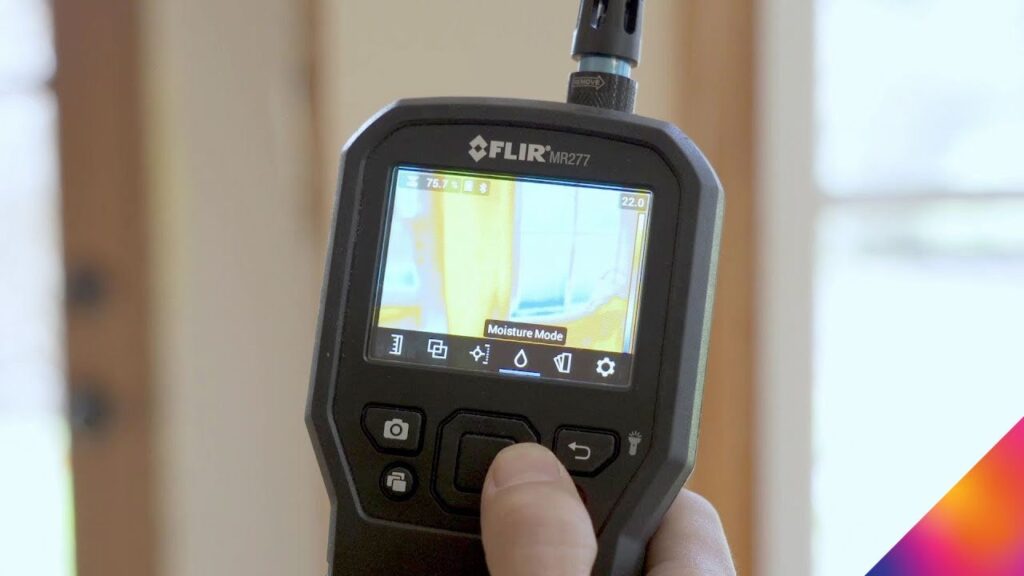 A hand holding a FLIR MR277 thermal moisture meter, displaying a moisture-detecting thermal image on its screen.