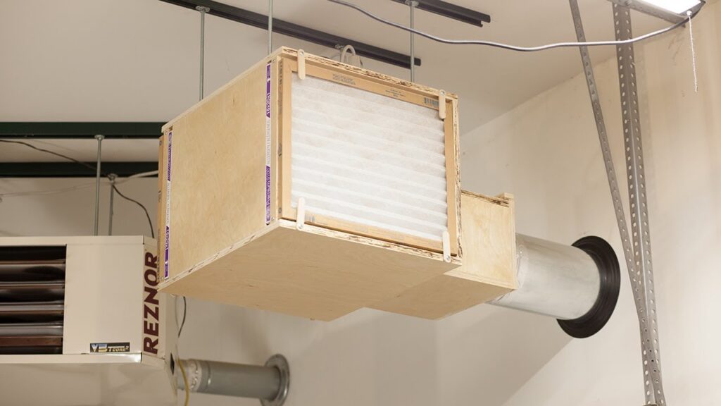 A wooden air filtration box with a white filter suspended from a ceiling next to a Reznor heater.
