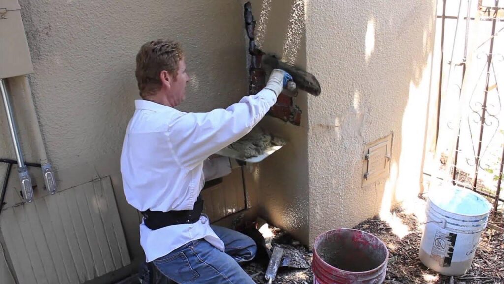 A man uses a trowel to apply stucco onto a damaged corner of an exterior wall.