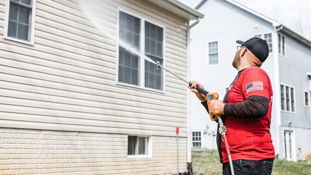 A man in a red shirt uses a pressure washer to clean the siding and windows of a light-colored house.