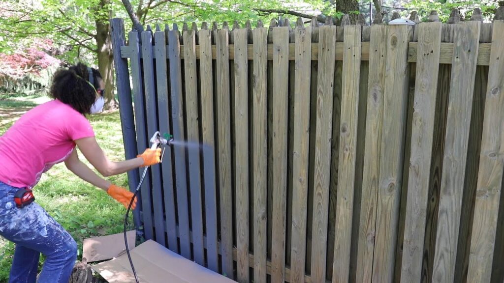 A woman wearing a pink shirt and protective gear uses a paint sprayer to paint a wooden picket fence dark gray.