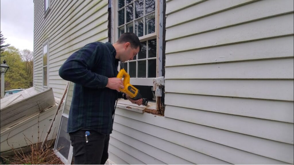 A man using a yellow reciprocating saw to cut the wooden frame around an exterior house window.