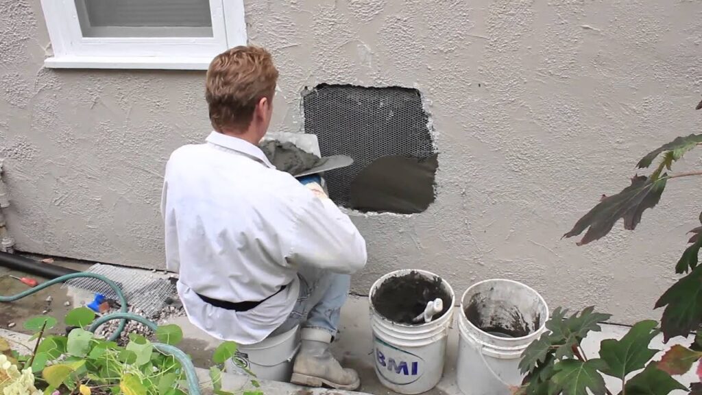 A worker applying wet mortar to a metal mesh patch on an exterior stucco wall.