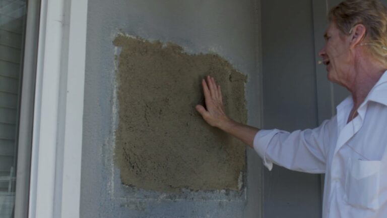 A man placing his hand flat against a newly patched square of stucco on a gray wall.