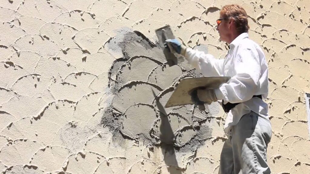 A plasterer uses a trowel to apply and texture fresh stucco on a beige wall.