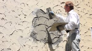 A plasterer uses a trowel to apply and texture fresh stucco on a beige wall.