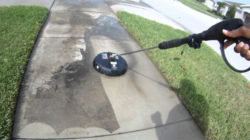 A person uses a pressure washer surface cleaner to clean a dirty concrete sidewalk.