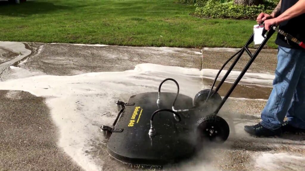 Person operating a wheeled surface cleaner to power wash a concrete driveway covered in white cleaning foam.