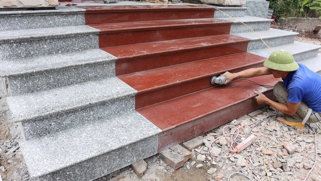 A worker in a green hat uses an angle grinder to polish red granite steps next to grey granite steps.