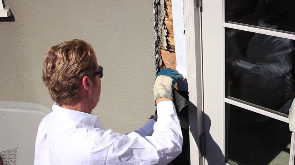 A construction worker in a white shirt and gloves installs waterproof flashing around the frame of a glass door.