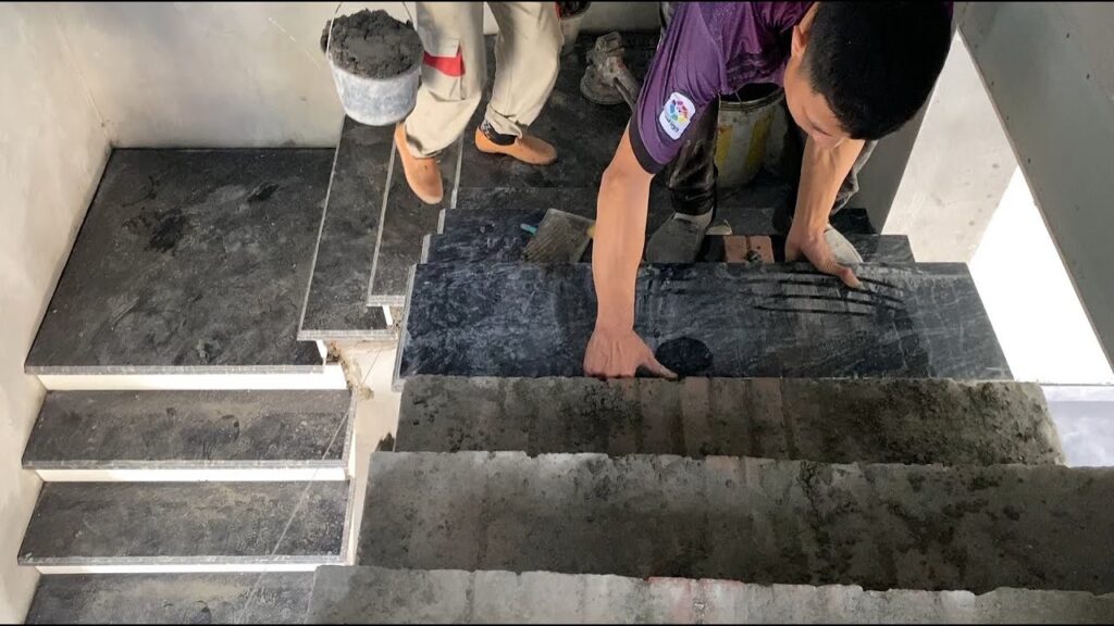 A construction worker installing black marble tiles on staircase steps with mortar.
