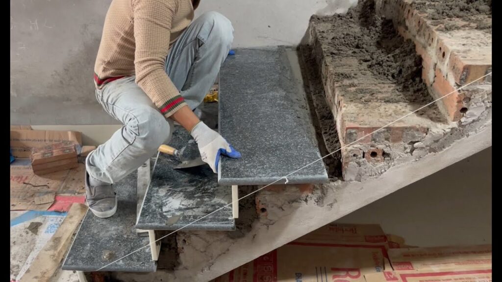 Worker wearing gloves installing a dark granite slab on concrete stairs.