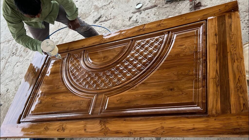 Worker spraying a glossy finish onto an intricately carved wooden door.