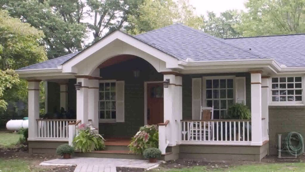 Front porch of a dark green house featuring white columns, a vaulted entryway, white railings, and potted plants.