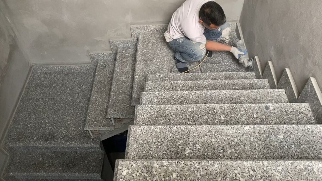 A construction worker uses a power grinder to polish grey terrazzo stairs.